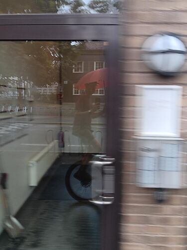 Picture of a reflection taken of a glass window. In the reflection is the person taking the picture. He is wearing a cap, t-shirt and shorts. He is holding his phone in one hand (to take the picture) and a red umbrella in the other. He is riding a unicycle.
