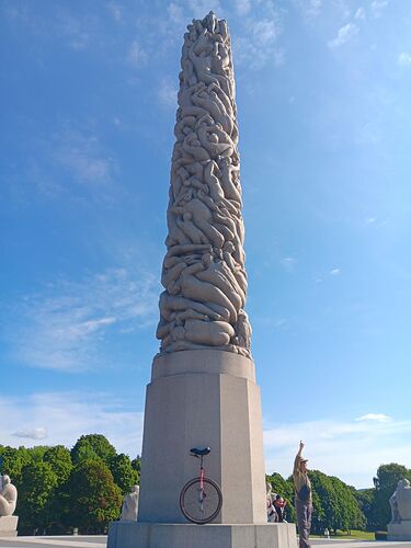 A red 29 inch wheeled unicycle in front of the Monolith scuplture (Monolitten). A 17 metre high carved stone column made up of naked human bodies of various ages and genders that is part of Frogner Park in Oslo.